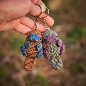 Puede incluir: Dos llaveros de ornitorrinco hechos a mano con ganchillo. Uno es marrón con detalles azules, el otro es gris con detalles morados. Ambos tienen llaveros. Los llaveros se sostienen contra un fondo verde borroso.