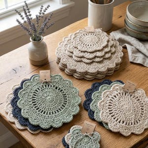 May include: A collection of handmade crochet coasters in various sizes and colors, including beige, green, and navy blue. The coasters have a scalloped edge and are arranged on a wooden table. A small vase with lavender and other kitchen items are also visible.