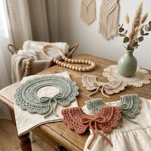 Puede incluir: Una colección de cuellos de ganchillo hechos a mano en varios colores, incluyendo verde salvia y terracota. Los cuellos se exhiben sobre una mesa de madera con un fondo de tonos neutros. También se ve un jarrón con flores secas y una cesta con mantas.
