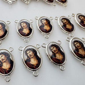 May include: A collection of oval-shaped silver-toned metal charms, each featuring a detailed portrait of Jesus wearing a crown of thorns. Each charm has three small circular loops for attachment, arranged in a grid pattern on a plain surface.