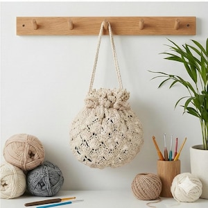 以下が含まれることがあります： 木製のペグラックに、テクスチャード加工のベージュのかぎ針編み巾着バッグが吊り下げられています。ニュートラルカラーの毛糸玉と、かぎ針編みが白い表面に配置されており、手作りやハンドメイドのテーマを示唆しています。