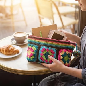 Op de afbeelding: Een kleurrijke, gehaakte etui met een regenboogritssluiting en pompomdetail. De etui heeft vierkante patronen in turquoise, roze en groen. Een croissant, koffie en een notitieboekje liggen op een tafel.