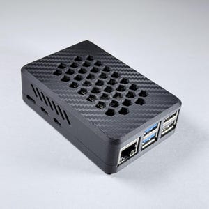 Puede incluir: Una carcasa electrónica rectangular negra con un patrón de fibra de carbono. La parte superior tiene un diseño de ventilación de panal. La parte frontal tiene varios puertos, incluyendo USB y Ethernet. La carcasa está diseñada para albergar componentes electrónicos.
