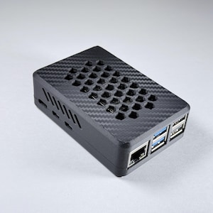 Puede incluir: Una carcasa electrónica rectangular negra con textura de fibra de carbono. La parte superior tiene un patrón de panal para ventilación. La parte frontal tiene varios puertos, incluyendo USB y Ethernet. La carcasa está diseñada para componentes electrónicos.