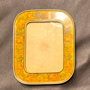 May include: A rectangular photo frame with rounded corners, featuring a floral pattern of yellow roses and green leaves on a gold background. The frame has a gold border and a clear glass or plastic insert.