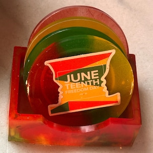 May include: A set of round, translucent resin coasters in red, yellow, and green, housed in a matching square holder. The front coaster features a graphic with the text JUNE TEENTH FREEDOM DAY, designed with a silhouette profile shape.