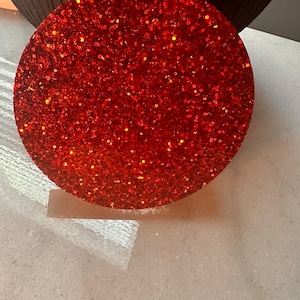 May include: A circular coaster featuring a vibrant red finish embedded with numerous fine glitter particles that create a reflective and textured appearance. The surface is uniform and round, showcasing a dense concentration of sparkling red specks.