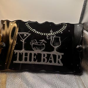Peut inclure: Un plateau rectangulaire en résine noire aux bords ondulés avec un accent latéral doré. La surface présente des graphismes à paillettes argentées de trois verres à cocktail et le texte THE BAR en lettres capitales, avec des cristaux incrustés.