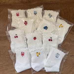 May include: A collection of white ribbed ankle socks with ruffled cuffs, each individually packaged in clear plastic. The socks feature small, colorful embroidered motifs including cherries, strawberries, blueberries, lemons, a lobster, and fish.