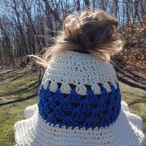 May include: A white and blue crochet hat with a messy bun hairstyle. The hat features a white top section and a blue patterned lower section. The hat is handmade and suitable for outdoor wear.