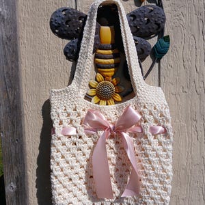 Pode incluir: Bolsa tote de crochê creme com um laço de cetim rosa. A bolsa tem um padrão texturizado e uma alça resistente. Uma abelha e um girassol decorativos de metal estão no fundo, presos a uma superfície de madeira.