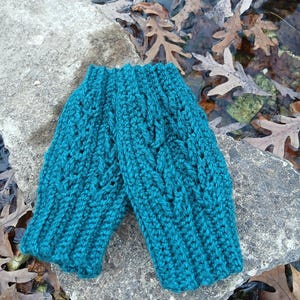 May include: Teal blue crocheted fingerless gloves with a cable knit pattern. The gloves are lying on a gray rock with brown leaves scattered around.