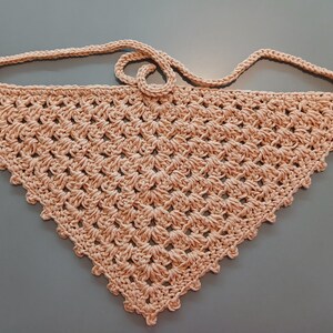 May include: A peach-colored, handmade crochet bandana with a triangular shape. The bandana features a textured, open weave design and a strap for tying around the neck. The edges are finished with a scalloped trim.