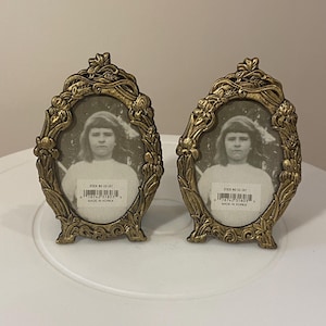 May include: Two antique gold-colored oval picture frames with a floral design. Each frame holds a black and white photograph of a person. The frames have a stand and are labeled "STOCK NO ES-247" and "MADE IN KOREA".