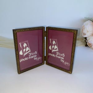 May include: A hinged, antique-style photo frame with a bronze-colored metal frame. Each side holds a 3.5x5 inch photo, with a burgundy background. The frame is open, displaying the interior with the text "3 1/2 x 5 photo frame by".