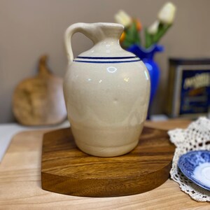 May include: A cream-colored ceramic jug with a single handle and two thin, horizontal blue stripes encircling the upper portion of the body. The jug features a smooth, rounded silhouette with a narrow neck and a slightly flared rim at the top.