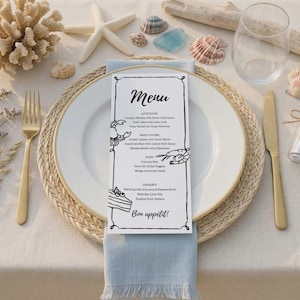 Pode incluir: Um cartão de menu com uma borda preta e a palavra "Menu" em itálico. O menu lista aperitivos, pratos principais, acompanhamentos e sobremesas. O cartão está em um prato branco com borda dourada, um guardanapo azul e talheres dourados. Conchas e uma estrela do mar estão espalhadas.