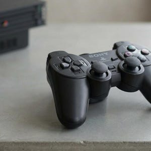 Peut inclure: Manette noire Sony PlayStation 2 avec plusieurs boutons et des joysticks analogiques. La manette est posée sur une surface grise, avec une console noire en arrière-plan. Le mot "SONY" est visible sur la manette.