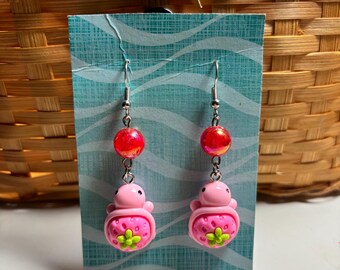 May include: A pair of drop earrings featuring a pink turtle-shaped charm with a strawberry-patterned shell, a small green flower accent, and a round, translucent red bead suspended from a silver-toned metal hook on a light blue display card.