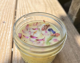May include: A small, clear glass jar candle filled with yellow wax and decorated with dried flower petals. The candle has a white wick and sits on a wooden surface. The petals are pink, purple, and white.
