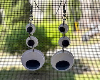 May include: A pair of quirky dangle earrings with a layered design. Each earring has three white circular elements with black oval accents, connected by silver-tone metal. The earrings hang from silver-tone ear wires, creating a playful, eye-catching accessory.
