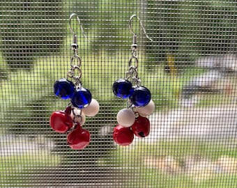 May include: A pair of drop earrings featuring silver-toned metal hooks and chains. Each earring is adorned with a cluster of small, round, glossy beads in solid red, white, and deep blue, suspended from the chain in a multi-layered arrangement.