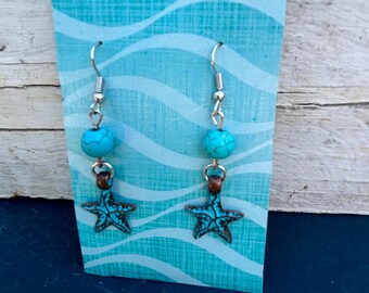 May include: Dangle earrings featuring turquoise beads and bronze starfish charms. The earrings are suspended from silver-coloured fish hooks. They are presented on a blue card with a wave pattern.