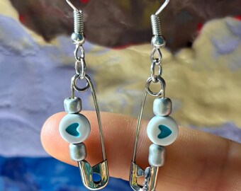 May include: Silver earrings with a safety pin design. Each earring has a silver hook, silver beads, and a white bead with a teal heart. The earrings are shown against a colourful background.