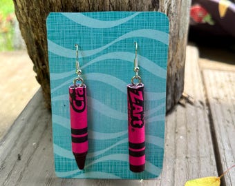 May include: Handmade earrings with a pink crayon design, black accents, and the text "Crayola" and "Zart". The earrings are displayed on a teal card with a wave pattern, against a wooden background.