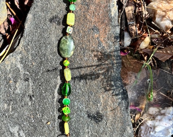 May include: A beaded suncatcher featuring green and yellow beads of various shapes, including oval, round, and square. A clear crystal prism hangs at the bottom. The beads are strung on a wire and are displayed against a grey stone.