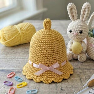 Peut inclure: Un chapeau en forme de cloche jaune crocheté avec un nœud en ruban rose. Le chapeau a un bord festonné et un petit bouton sur le dessus. Un lapin crocheté tenant un œuf est en arrière-plan, avec une pelote de laine jaune et des marqueurs de point colorés.