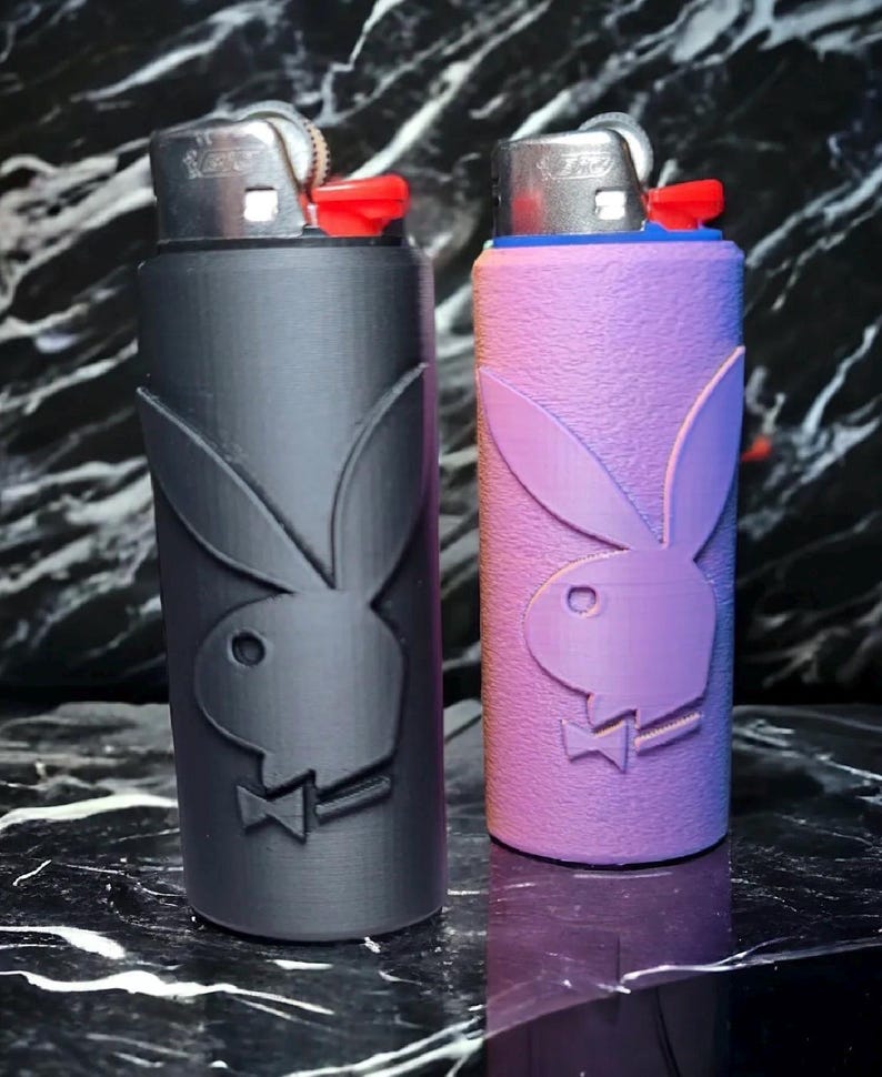 Puede incluir: Dos encendedores con fundas personalizadas. Uno es negro, el otro es morado. Ambas fundas presentan el logotipo del conejo Playboy. Los encendedores tienen detalles en rojo y azul.