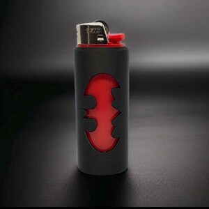 Puede incluir: Encendedor negro y rojo con un dise&ntilde;o del logotipo de Batman. El encendedor tiene un cuerpo negro con un s&iacute;mbolo de murci&eacute;lago rojo recortado, revelando el interior rojo. La parte superior es plateada con un bot&oacute;n de encendido rojo.