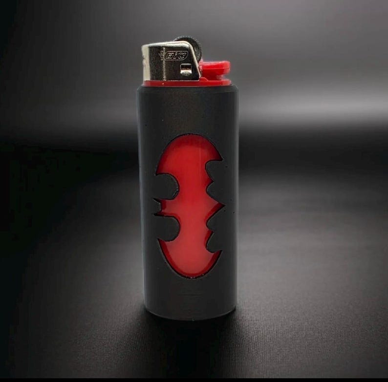 Puede incluir: Encendedor negro y rojo con un dise&ntilde;o del logotipo de Batman. El encendedor tiene un cuerpo negro con un s&iacute;mbolo de murci&eacute;lago rojo recortado, revelando el interior rojo. La parte superior es plateada con un bot&oacute;n de encendido rojo.