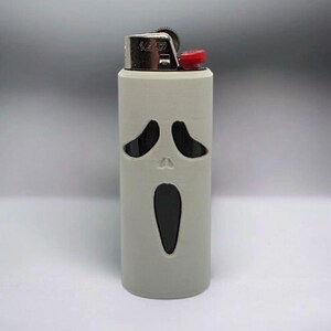 Puede incluir: Una funda de encendedor gris con un dise&ntilde;o recortado de una cara de fantasma gritando. El encendedor tiene una parte superior plateada con un bot&oacute;n rojo. El dise&ntilde;o est&aacute; inspirado en la franquicia de pel&iacute;culas de terror.