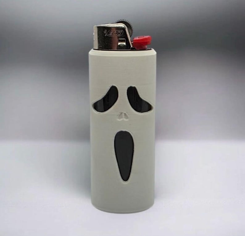 Puede incluir: Una funda de encendedor gris con un dise&ntilde;o recortado de una cara de fantasma gritando. El encendedor tiene una parte superior plateada con un bot&oacute;n rojo. El dise&ntilde;o est&aacute; inspirado en la franquicia de pel&iacute;culas de terror.