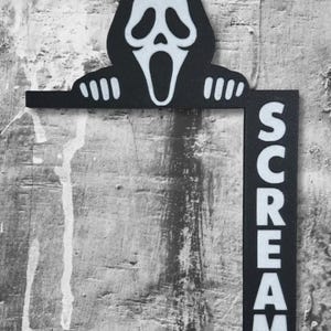 May include: A black and white wall-mounted sign featuring the iconic Ghostface mask character with hands resting on a horizontal bar. A vertical rectangular section attached to the right side displays the word SCREAM in bold white capital letters.