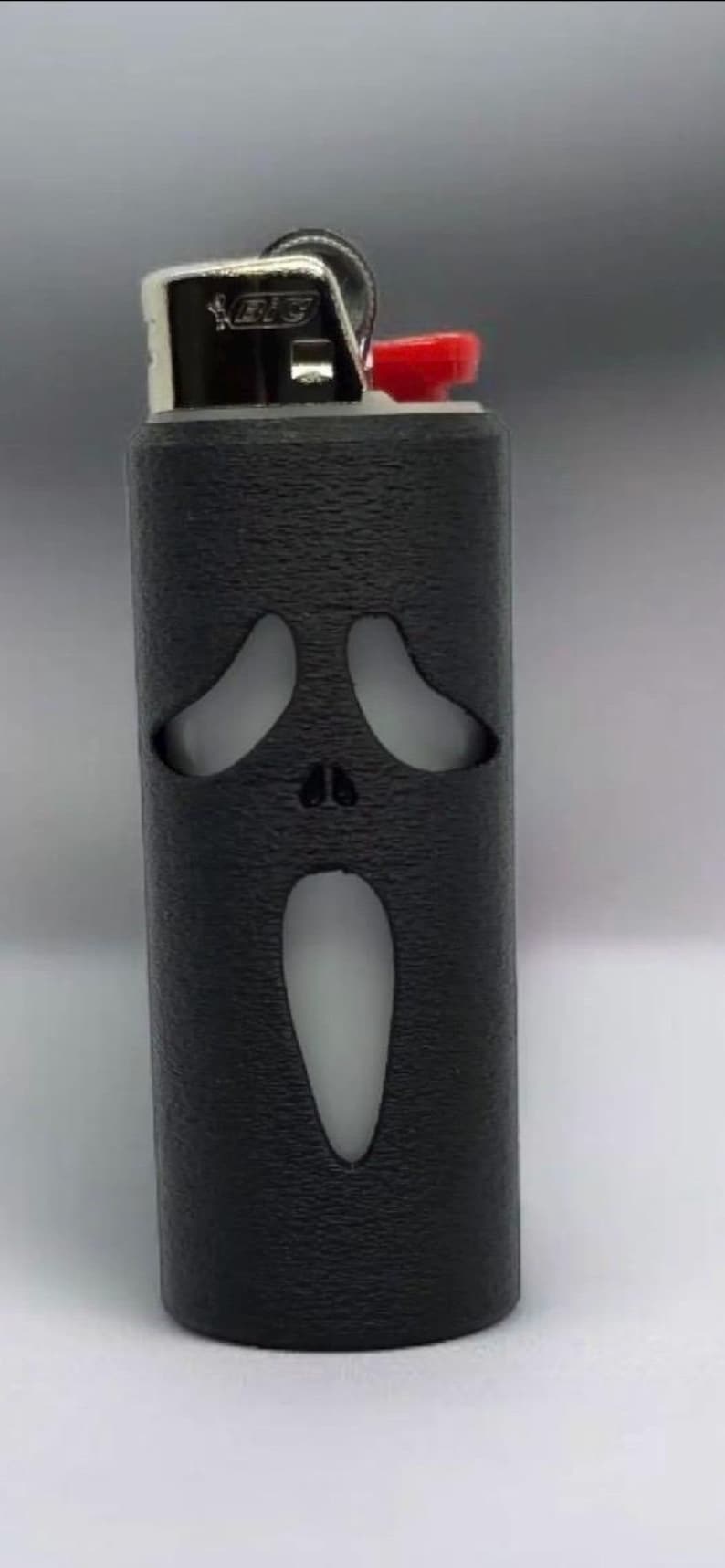 Puede incluir: Un encendedor negro con una superficie texturizada, con un dise&ntilde;o recortado de la m&aacute;scara de Ghostface de la franquicia Scream. El encendedor tiene un bot&oacute;n de encendido rojo y una parte superior de metal plateado.