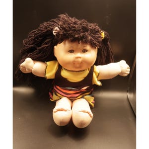 May include: A vintage doll with a round face, brown yarn hair, and a yellow shirt under a brown jumper. The doll's skirt features colorful stripes. The doll has open arms and is sitting down.