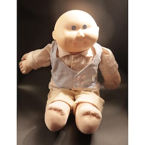 May include: A vintage Cabbage Patch Kid doll with a bald head and blue eyes. The doll is wearing a light tan button-down shirt, a light blue vest, and tan pants. The doll's arms and legs are outstretched.