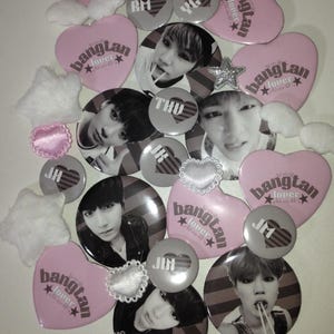 May include: A collection of circular and heart-shaped pin-back buttons featuring black and white portraits of individuals, striped patterns, and pink heart-shaped buttons with the text bangtan lover. Includes small white plush stars and wings.