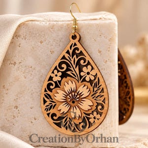 May include: A teardrop-shaped wooden earring featuring an intricate laser-cut floral pattern with a central flower and surrounding leaves. The earring hangs from a gold-toned metal hook. The text CreationByOrhan is visible at the bottom.