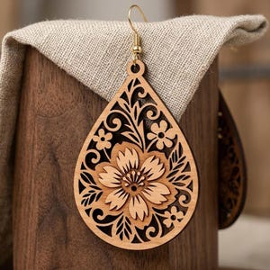 May include: Wooden teardrop earrings with a floral design. The earrings are light brown with a laser-cut floral pattern. The earrings have gold-coloured ear wires and are displayed on a wooden block with a beige cloth.
