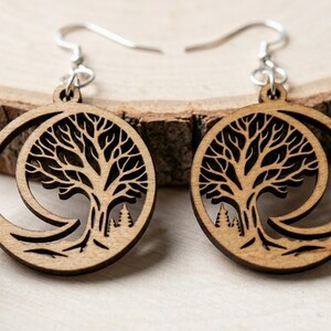 May include: A pair of wooden earrings featuring a tree of life design. The earrings are circular with a cutout tree and crescent moon shape. The earrings have silver-colored hooks. The earrings are made of light brown wood.