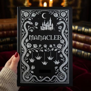Peut inclure: Un livre noir avec le mot "MANACLED" en blanc. La couverture présente une illustration blanche d'un château, de roses, de serpents et de grues en origami. Le livre est tenu par une personne avec du vernis à ongles rouge.