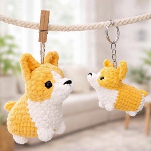 May include: Two crochet corgi dog ornaments, one hanging from a wooden clothespin and the other attached to a keychain. The dogs are yellow and white, with black eyes and noses. The background is blurred.