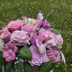 Può includere: Un'arrangiamento floreale di sapone nei toni del rosa, viola e bianco. Include rose, dalie e altre forme di fiori, accentuato da perle finte e foglie verdi. Un fiocco decorativo aggiunge un tocco finale.