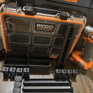 May include: An open black and orange RIDGID Pro Gear System Gen 2.0 tool case with various black tool organizers and an orange and black drill. The case is clear with a black grid design and the RIDGID logo.