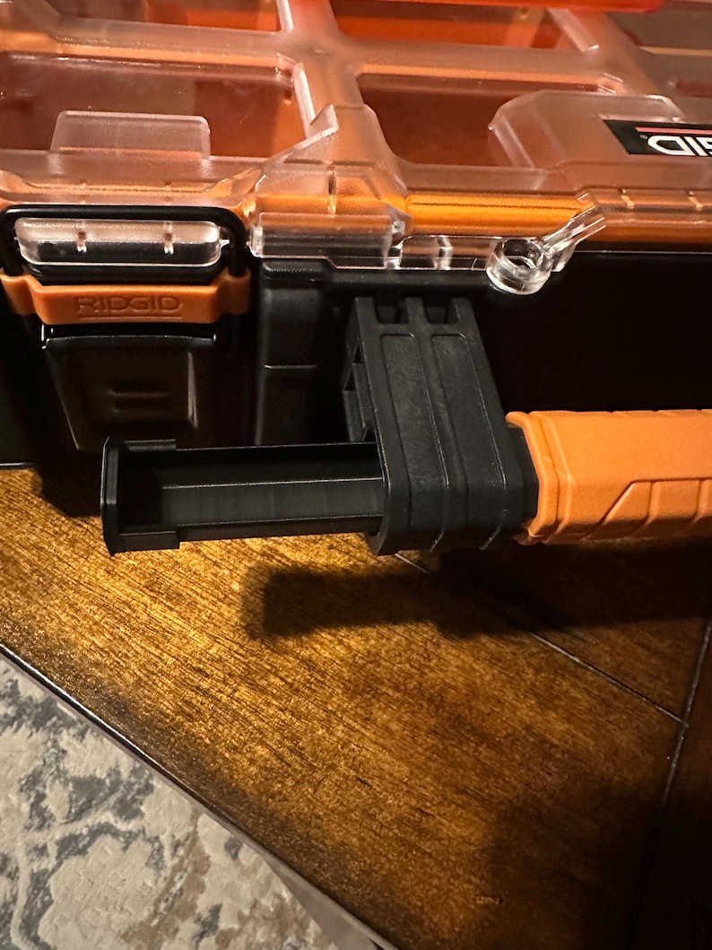 May include: Close-up of a black and orange RIDGID tool organizer. The organizer has a clear lid, orange accents, and a pull-out drawer. The RIDGID logo is visible on the front. The organizer is on a wooden surface.