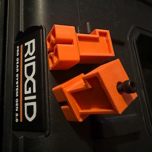 May include: Two bright orange, 3D-printed components with a rectangular design and interlocking features. One component has a black bolt. The items are on a black surface, near a label that reads "RIDGID PRO GEAR SYSTEM GEN 2.0".